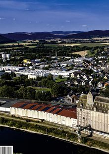 Stadtansicht mit Häusern, Feldern und Hügeln im Hintergrund, ein Fluss im Vordergrund, Fabrikgebäude mit rotem Dach.