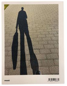 Unten links steht "maat". Ein langer Schatten einer Person mit einer Tasche auf einem gepflasterten Gehweg.