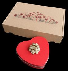 Ein brauner Karton mit Geschenkmuster, davor eine herzförmige rote Schachtel mit goldener Schleife.