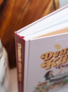 Ein Buch mit rotem Rücken zeigt den Titel "Deutschlands bester Koch" und "ZUCKER". Ein Glas steht daneben.
