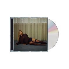 Eine Frau mit langem, hellem Haar liegt in dunklem Pulli und schwarzer Strumpfhose auf dem Boden vor einem Fenster mit Vorhängen.