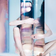 Person umarmt zwei große Glaszylinder, Reflektionen und Farbmuster verzerren den Körper. Hände tragen Ringe, ruhige Atmosphäre.