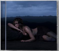 Eine Frau liegt in dunkler Abendlandschaft auf sandigem Boden, umgeben von Bergen unter einem bewölkten Himmel.