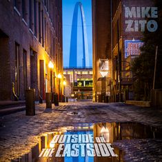 MIKE ZITO, OUTSIDE OR THE EASTSIDE; Pfad zwischen Gebäuden, beleuchtet, im Hintergrund ist ein großer Bogen.