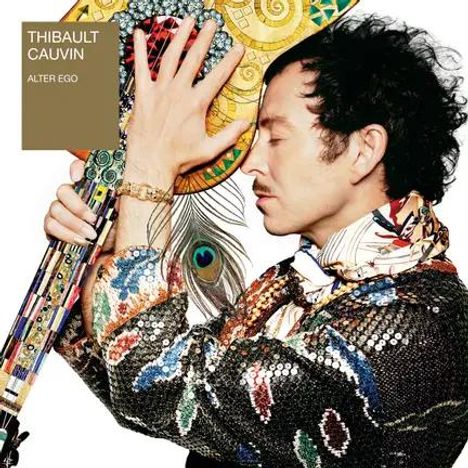 Text: "THIBAULT CAUVIN ALTER EGO". Ein Mann hält ein buntes Objekt, trägt ein glitzerndes Outfit und ein buntes Halstuch.
