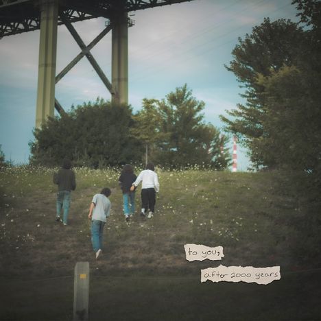 Text: "to you, after 2000 years." Vier Menschen gehen einen grasbewachsenen Hügel hinauf, im Hintergrund eine Brücke.