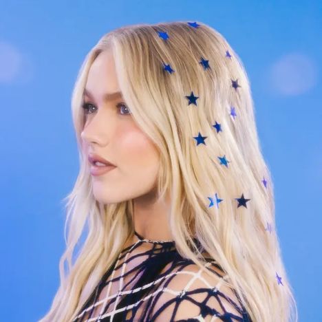 Keine erkennbaren Texte. Eine Frau mit blonden Haaren, darin blaue Sternensticker. Hintergrund in hellblau.