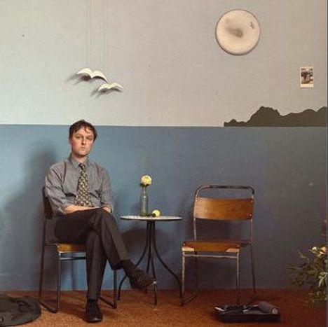 Ein Mann sitzt auf einem Stuhl neben einem kleinen Tisch mit einer Blume. An der Wand hängen Vögel und ein Bild.