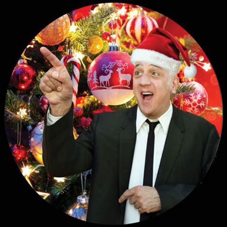 Ein Mann in Anzug und Weihnachtsmütze zeigt lachend auf einen geschmückten Weihnachtsbaum mit roten Kugeln.