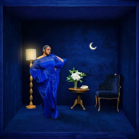 Eine Frau in blauer Kleidung steht in einem blauen Raum mit Stehlampe, Tisch mit Blumen, Stuhl und einer Mondsichel an der Wand.