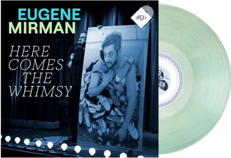 Text links: "Eugene Mirman", "Here Comes The Whimsy". Darunter eine Person mit einem Plakat, im Hintergrund Vorhang. Rechts: transparente Schallplatte.