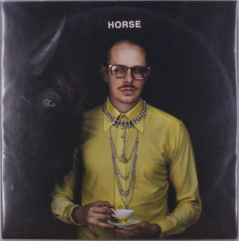 Der Text "HORSE" steht oben. Ein Mann in gelber Bluse mit Brille hält eine Teetasse, neben ihm ein dunkles Pferd.