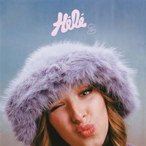 Text: „Héla 2“. Eine Frau mit lila Flauschhut zieht ein witziges Gesicht vor einem dezenten Hintergrund.