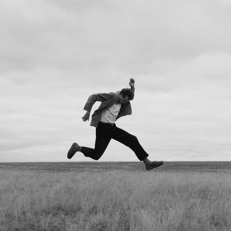 Kein Text vorhanden. Ein Mann springt dynamisch in die Luft über ein Grasfeld, Himmel wolkig.