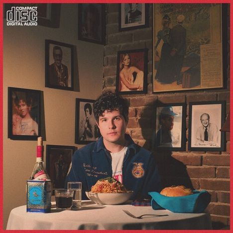 „COMPACT DISC DIGITAL AUDIO“ oben links. Ein Mann sitzt an einem Tisch mit Essen. Umgeben von zahlreichen Fotos an der Wand.