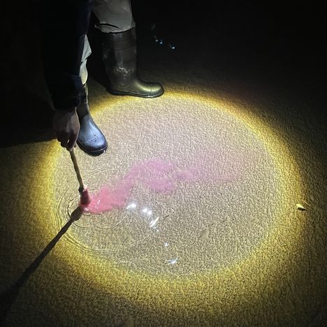 Ein stiefeltragender Mensch leuchtet auf eine Pfütze, in die er mit einem Stab pinke Farbe rührt.