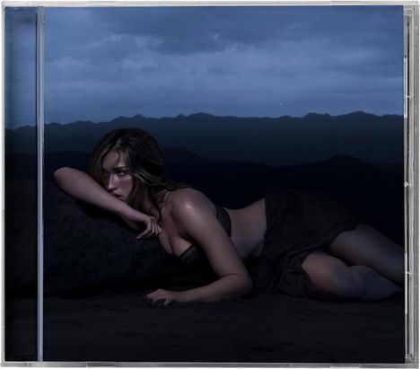 Eine Frau liegt in dunkler Abendlandschaft auf sandigem Boden, umgeben von Bergen unter einem bewölkten Himmel.