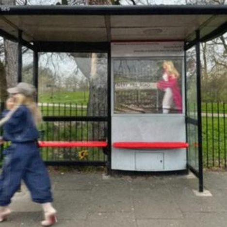 Ein Wartehäuschen mit einer Frau im Vordergrund. Klare Struktur, leuchtend roter Sitz. Hintergund: Park und Bäume.