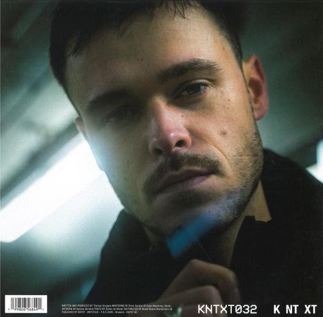 KNTXT032 K NT XT. Ein Mann schaut ernst in die Kamera, im Hintergrund unscharfes Licht.