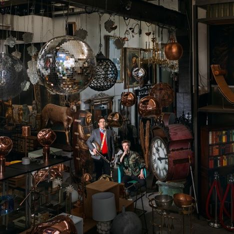 Zwei Personen in einem vollgestellten Raum mit Spiegelkugeln, antiken Uhren, Kupferlampen und einem Hirschkopf.