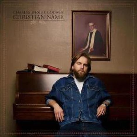 Text: "Charles Wesley Godwin Christian Name". Ein Mann in Jeansjacke sitzt vor einem Klavier, darüber ein Porträt an der Wand.
