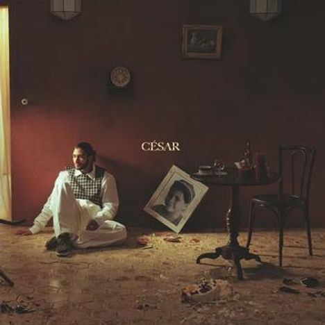 Der Text "CÉSAR" steht zentral. Ein Mann sitzt auf dem Boden, daneben ein Tisch mit Wein und ein schiefes Porträt.