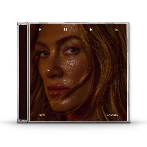 Text: PURE, DELTA, GOODREM. Porträt einer Frau mit glänzendem Gesicht und brünettem Haar, Nahaufnahme.