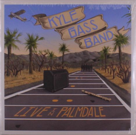 "Kyle Gass Band" und "Live in Palmdale" auf einer Straße. Flugzeug mit Banner und Wüstenlandschaft mit Palmen im Hintergrund.