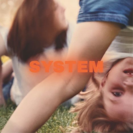 Text "SYSTEM" in orange. Unscharfes Bild von Personen auf Gras, ein Kind im Vordergrund, teilweise kopfüber.