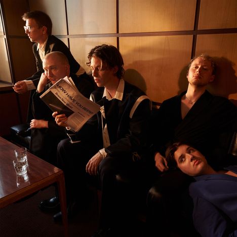 "HOW HAVE YOU BEEN?" auf Zeitung, fünf Männer in formeller Kleidung in einem Raum mit Holzwänden; zwei Gläser auf Tisch.