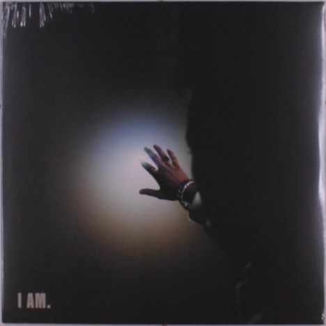 Der Text „I AM.“ ist weiß auf schwarzem Hintergrund. Eine Hand, geschmückt mit Schmuck, greift ins Licht.