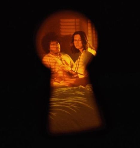 Zwei Menschen sind durch ein Schlüsselloch zu sehen, in warmes Licht getaucht, sitzen auf einem Bett.