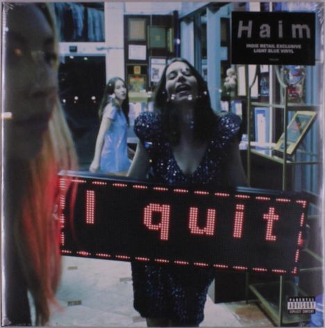 Text: "Haim", "I quit". Junge Frau in schimmerndem Kleid hält ein Schild nachts vor einem Laden.