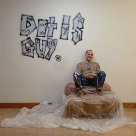 Text: "DAT IS GUY!" in graffiti-Stil. Eine Person sitzt auf einem Plastik-bedeckten Sessel und lächelt, in einem hellen Raum.