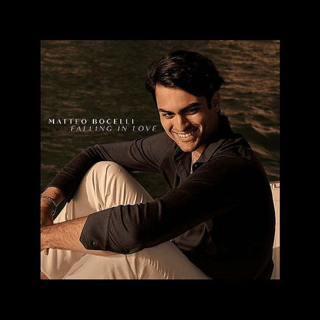 MATTEO BOCELLI, FALLING IN LOVE. Ein Mann lächelt, sitzt entspannt am Wasser in einem dunklen Hemd und hellen Hosen.