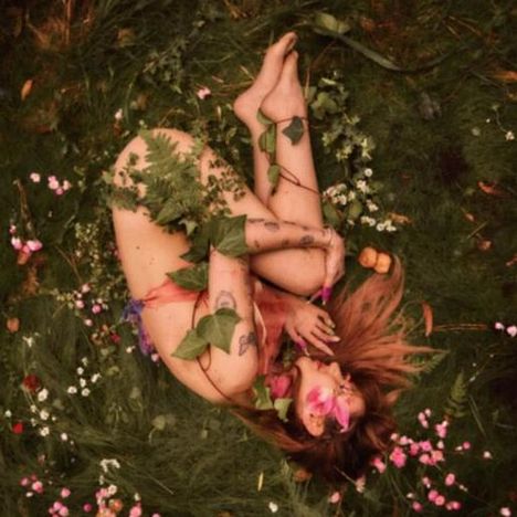 Eine Person liegt nackt in einem Bett aus Gras und Blumen, mit Pflanzen bedeckt.