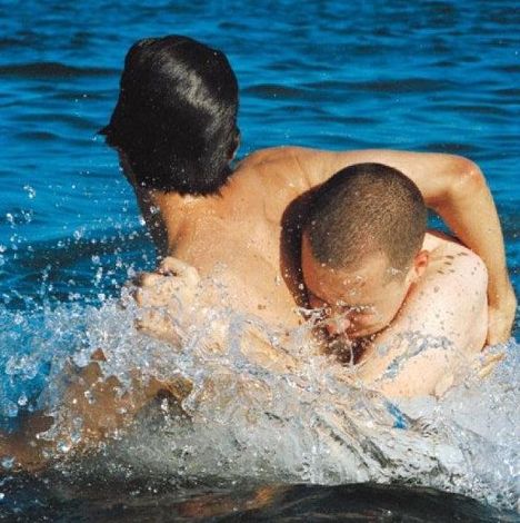 Zwei Männer umarmen sich im Wasser, Wellen spritzen um sie herum, die Szene wirkt intensiv und dynamisch.