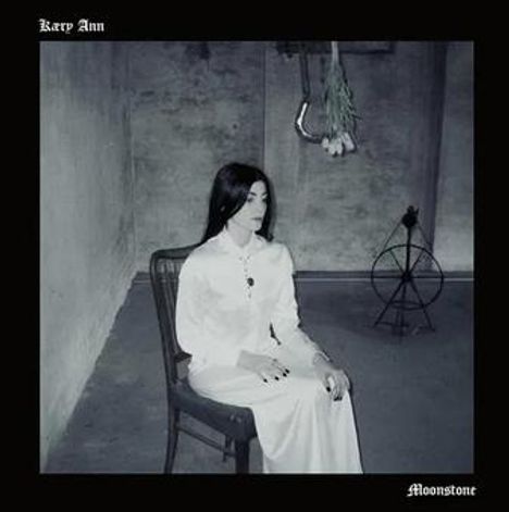 „Karp Ann“ oben links, „Moonstone“ unten rechts. Eine Frau in Weiß sitzt in einem kargen, grauen Raum.