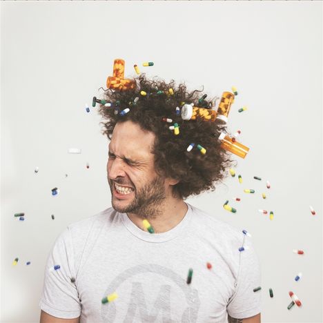 Ein Mann mit lockigem Haar, Augen geschlossen, von herumfliegenden Pillen umgeben, trägt ein T-Shirt mit einem stilisierten "M".