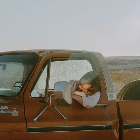 Ein rostiger Pickup mit Text "Custom Deluxe 10". Eine Person lehnt entspannt aus dem Fenster. Steinige Landschaft im Hintergrund.