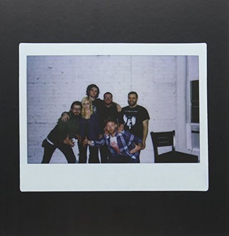 Eine Gruppe von sechs lächelnden Personen posiert vor einer weißen Backsteinwand. Eine leere schwarze Stuhllehne rechts.