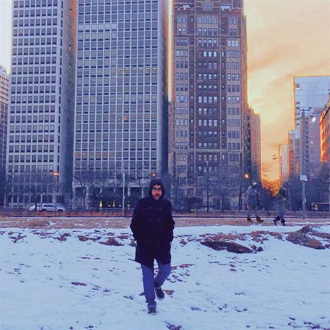 Person in dunkler Kleidung geht durch schneebedeckte Fläche, im Hintergrund sind hohe Gebäude bei Sonnenuntergang zu sehen.