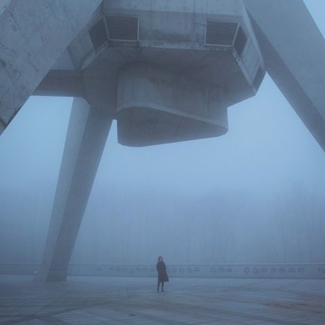 Überdimensionale Betonstruktur in nebliger Szenerie, eine Person steht allein darunter, umgeben von Bäumen im Hintergrund.