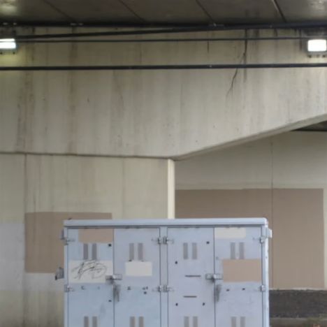 Graffiti auf einem grauen, metallischen Verteilerkasten unter einer Betonstruktur.