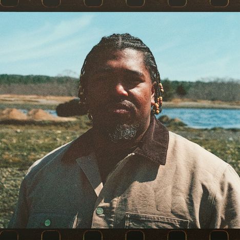 Keine sichtbaren Texte. Ein Mann mit geflochtenem Haar steht draußen vor einem Fluss bei klarem Himmel.