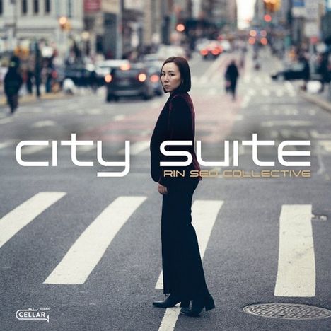 "City Suite", "Rin Seto Collective". Frau in dunkler Kleidung steht auf einem Zebrastreifen in einer belebten Stadtumgebung.