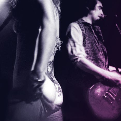 Zwei Personen, verschwommen. Eine zeigt den Rücken mit einem Tattoo; die andere spielt Gitarre, verschwommen beleuchtet.