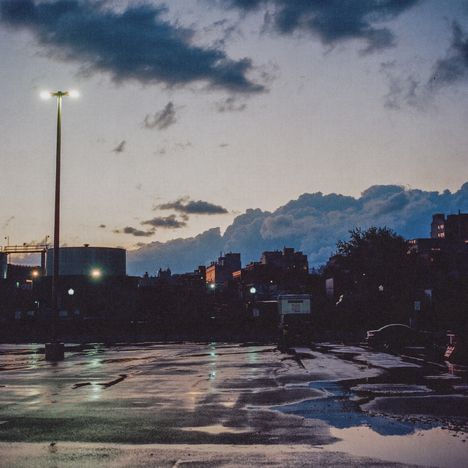 Ein leerer Parkplatz bei Dämmerung mit nasser Asphaltoberfläche. Im Hintergrund sind Gebäude und Wolken am Himmel zu sehen.