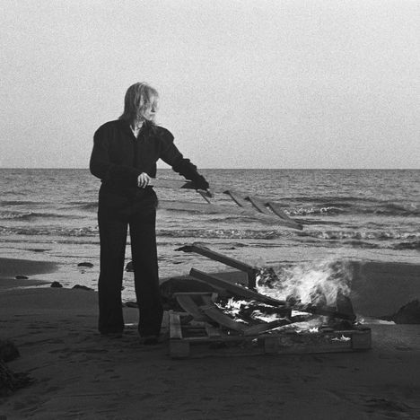 Eine Person steht am Strand, hält ein langes Objekt und bewegt es über ein knisterndes Feuer. Im Hintergrund das Meer.