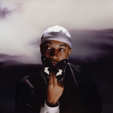 Ein Mann mit weißer Cap hält einen schwarzen Schmetterling mit weißen Flecken vor seinem Gesicht. Hintergrund scheint wolkig.